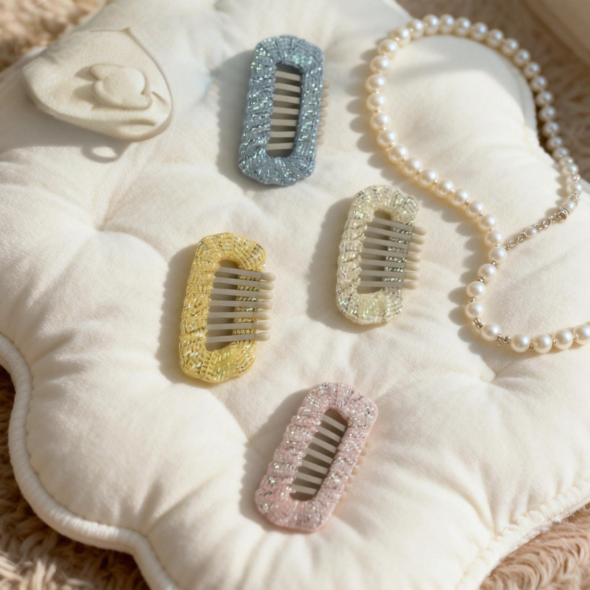 Aurora Weave Dog Barrettes Party Set. A flat lay of4-colors (Opal Blue, Butter Glow, Pearl Cream, Petal Rose) barrettes with gold sequin detail and a delicate pearl necklace, arranged on a light quilted mat. Perfect for creating a dazzling party look for small long-haired breeds.