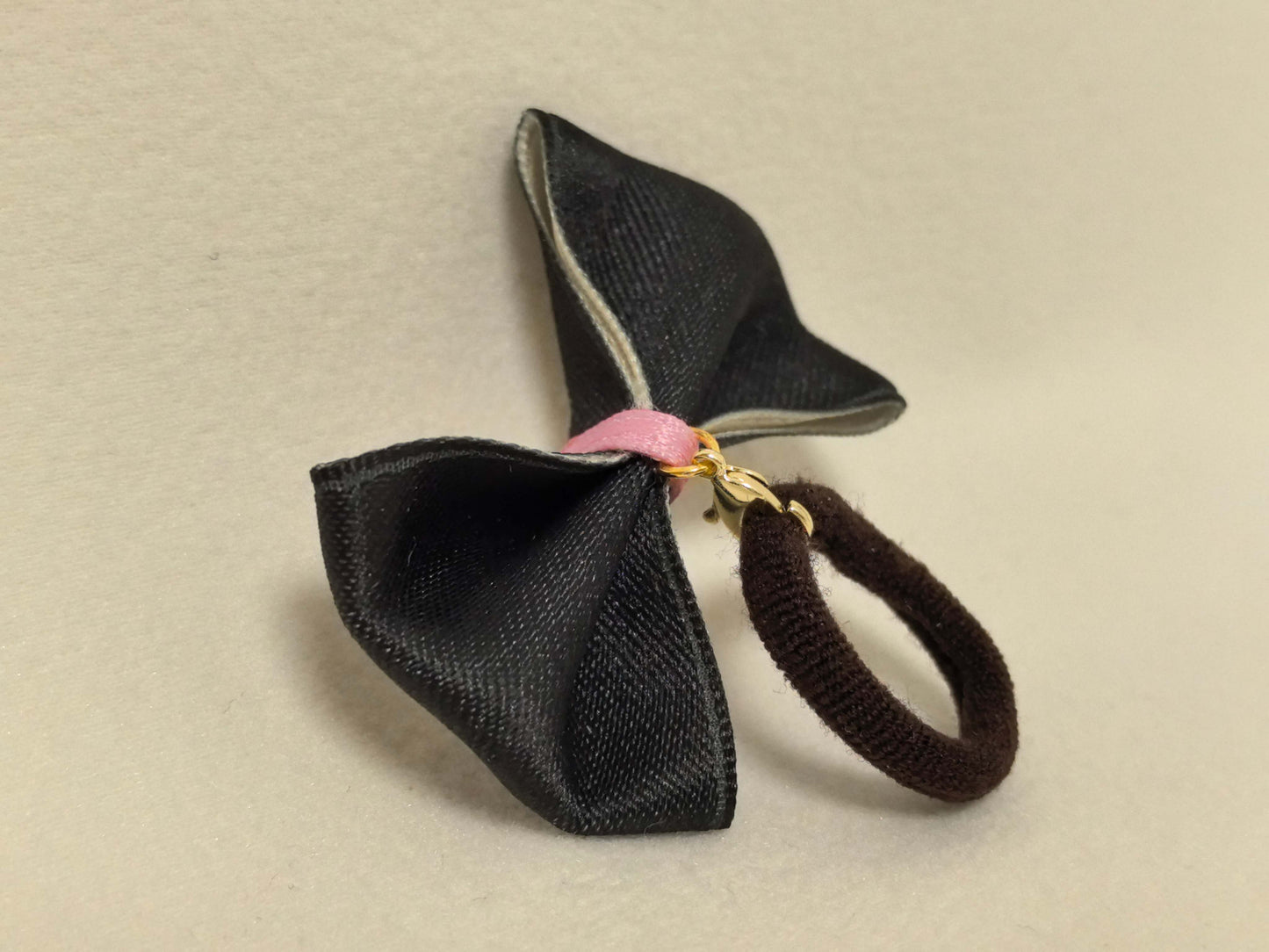 Side view of a black pet bow with a pink center, gold clasp, and attached dark brown hair tie. SilkyCo set.