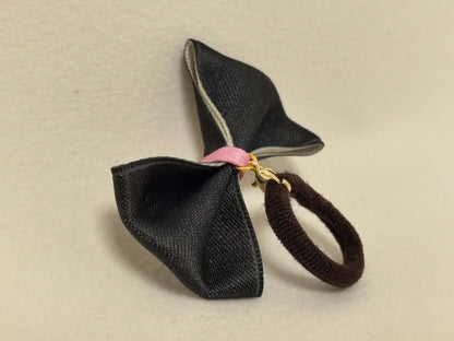 Side view of a black pet bow with a pink center, gold clasp, and attached dark brown hair tie. SilkyCo set.